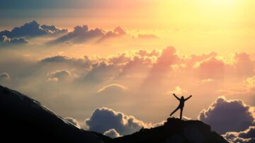 شخص يقف على قمة جبل مع انتشار الغيوم حوله، بينما تشرق الشمس في الأفق، مما يخلق مشهداً ساحراً وأجواء ملهمة.