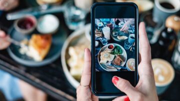 صورة تُظهر شخصًا يلتقط صورة لطبق طعام باستخدام هاتفه المحمول، مع وجود أطباق وقهوة على الطاولة. تُبرز الصورة الأجواء الاجتماعية لتناول الإفطار أو الغداء.