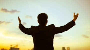 رجل يرتدي بدلة سوداء يقف في الهواء الطلق مع ذراعيه مرفوعتين، يواجه أفق المدينة تحت سماء مشمسة مع غيوم خفيفة. يعبر عن الإيجابية والتفاؤل.