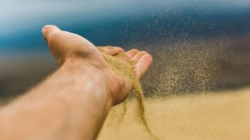 يد مفتوحة تحمل رمالًا تتساقط بشكل خفيف، مع خلفية ضبابية تجمع بين السماء والأرض. تعبر الصورة عن مفهوم الزمن والحرية، وتسلط الضوء على تفاصيل الرمال المتناثرة.