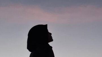 شخص يرتدي حجابًا أسود، يظهر في صورة ظلية ضد سماء ملونة بألوان دافئة، مما يبرز ملامح الوجه بشكل جميل.