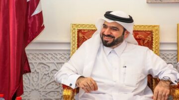 رجل يرتدي الزي التقليدي القطري يجلس مبتسمًا على كرسي مزخرف، خلفه علم قطر. يعكس الجو شعورًا بالترحيب والضيافة.