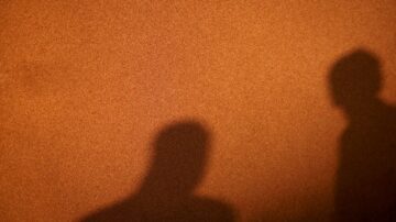 ظل شخصين يتشكلان على جدار بلون بني داكن، مع تباين واضح بين الظلال واللون. تعكس الصورة جوًا من الغموض والتفاعل.