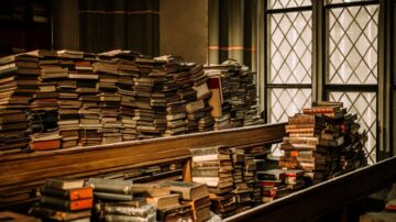 كتب قديمة مكدسة في مكتبة، مع نوافذ زجاجية تعكس الضوء. الكتب متنوعة في الأحجام والألوان، والبعض منها مغطى بالغبار، مما يبرز تاريخ المكان وعراقته.