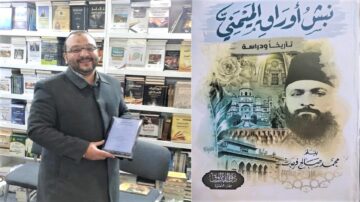 رجل مبتسم يحمل جائزة في يده، يقف أمام رفوف مليئة بالكتب. على الجانب الأيمن، غلاف كتاب بعنوان "تاريخ وأدب المدينة"، يظهر فيه صورة تاريخية لرجل يرتدي عمامة.