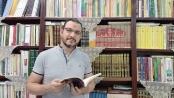 رجل يحمل كتابًا ويقف أمام رفوف مليئة بالكتب الإسلامية. تظهر تفاصيل زخرفية على الرفوف. تُبرز الصورة اهتمامه بالقراءة والثقافة.