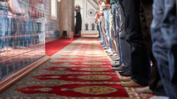 مجموعة من الأشخاص يقفون في صف للصلاة داخل مسجد، تظهر أرجلهم على سجادة مزخرفة، مع خلفية جدران مكسوة بالزخارف الإسلامية.