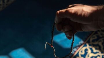 يد تحمل سبحة على خلفية سجادة زرقاء مزخرفة، مع إضاءة ناعمة تبرز تفاصيل اليد والسبحة.