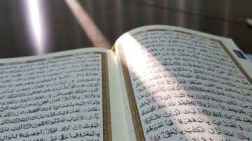 صفحة مفتوحة من القرآن الكريم مع أشعة الشمس تتسلل عبر الغلاف، تُبرز النص العربي المكتوب بخط جميل.