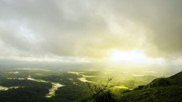 منظر طبيعي رائع يظهر واديًا أخضر تحت سماء غائمة مع أشعة الشمس تتسلل عبر السحب. تبرز المزارع في الأسفل، مما يخلق تباينًا جميلًا بين الطبيعة والمناطق الزراعية.