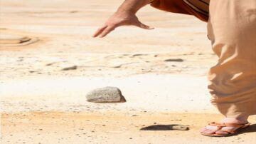 رجل يرتدي ملابس فضفاضة يقف على أرض صحراوية ويقوم بإلقاء حجر صغير بيده. تظهر تفاصيل التربة الجافة والأحجار في الخلفية، مما يعكس بيئة طبيعية قاسية.