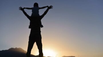 رجل يحمل طفلاً على كتفيه silhouetted against a sunset backdrop with mountains, symbolizing joy and family bonding.