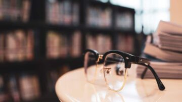 نظارات على طاولة بجانب مجموعة من الكتب في مكتبة. الخلفية تحتوي على رفوف مليئة بالكتب.