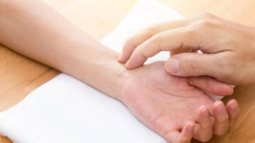 يد شخص تلمس معصم شخص آخر، مع التركيز على تقنيات الفحص الصحي. الخلفية خشبية، مع وجود منشفة بيضاء تحت اليدين.