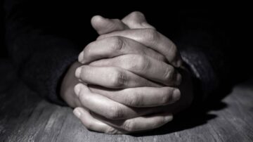 يد متشابكة في وضع تفكير، مع إضاءة خافتة تبرز تفاصيل الأصابع واليدين، خلفية داكنة تضيف عمقاً للصورة.