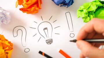 رسم توضيحي يظهر يدًا تكتب على ورقة بيضاء مع مصباح يضيء ورموز استفهام وعلامات تعجب. حول الورقة كرات ملونة من الورق الممزق. يعكس التصميم أفكارًا وإبداعًا.