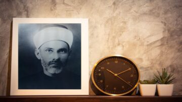 صورة تاريخية لرجل يرتدي عمامة بيضاء، مؤطرة بإطار أبيض، موضوعة بجانب ساعة حائط داكنة ونباتات صغيرة في أصيصين. خلفية الصورة جدارية بتصميم خشن.