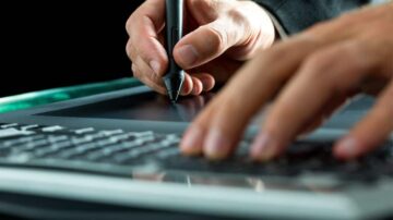 يد تُمسك قلمًا رقميًا وتكتب على لوح رسم رقمي مع وجود لوحة مفاتيح في الأسفل، تعكس الإبداع والتكنولوجيا الحديثة في التصميم الجرافيكي.