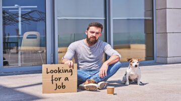رجل يجلس على الأرض بجانب كلبه، يحمل لافتة مكتوب عليها "أبحث عن عمل"، في بيئة حضرية مع نوافذ زجاجية في الخلفية. يعكس المشهد شعورًا بالبحث عن فرصة عمل ورغبة في الدعم.