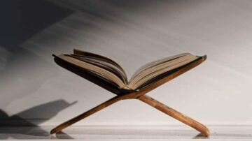 كتاب مفتوح على حامل خشبي، يظهر من زاوية جانبية مع ظلال ناعمة، يبرز تفاصيل الصفحات والأغلفة.