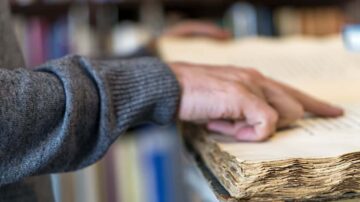 يد تظهر وهي تلمس صفحات كتاب قديم، مع تركيز على تفاصيل الصفحات الممزقة والمتهالكة. خلفية غير واضحة تحتوي على رفوف كتب.