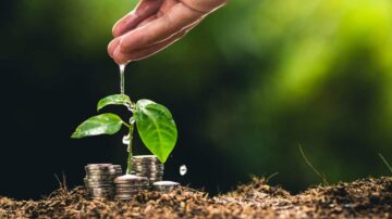 يد تروي نبتة صغيرة تنمو بين أكوام من العملات المعدنية، مما يرمز إلى الاستثمار المستدام والنمو المالي. الخضرة تعكس الأمل والازدهار، بينما تعبر القطرات المتساقطة عن العناية والرعاية في عالم المال.