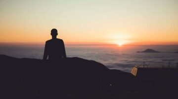 رجل يقف silhouetted أمام غروب الشمس فوق السحب، مع مشهد جبلي في الخلفية، يعكس جمال الطبيعة وهدوء اللحظة.