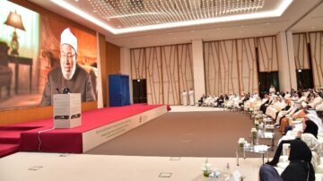 اجتماع رسمي في قاعة حديثة مع جمهور كبير، حيث يظهر متحدث على شاشة كبيرة. يتضمن المشهد رجال ونساء يرتدون ملابس تقليدية، ويجلسون في صفوف مرتبة. الإضاءة الساطعة والتصميم العصري يضيفان أجواء مهنية للحدث، الذي يبدو أنه يتعلق بمناقشات دينية أو ثقافية.