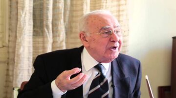 رجل مسن يرتدي بدلة رسمية يتحدث بحماس، مع تعابير وجه تعكس الشغف. خلفية غرفة ذات ستائر بيج، تضفي طابعًا دافئًا.