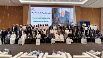 مجموعة من المشاركين في مؤتمر دولي، يجلسون في قاعة كبيرة. خلفهم شاشة تعرض صورة لشخصية مهمة مع نص باللغة العربية. الحضور يتضمن رجال ونساء من مختلف الجنسيات، يرتدون ملابس رسمية.