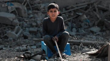 طفل يجلس على صندوق كرتوني في منطقة مدمرة، محاط بأنقاض وأشياء مكسورة، يبدو عليه القلق.