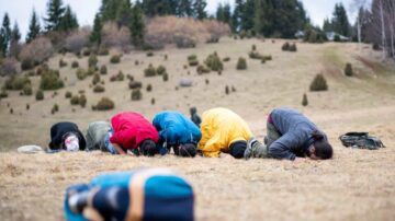 مجموعة من الأشخاص يؤدون السجود في منطقة عشبية، يرتدون ملابس ملونة، مع أشجار في الخلفية. تعكس الصورة روح العبادة والسكينة في الطبيعة.