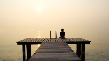 رجل يجلس على رصيف خشبي يطل على بحيرة هادئة في ضوء شمس الغروب، مع أجواء ضبابية تعكس جمال الطبيعة.