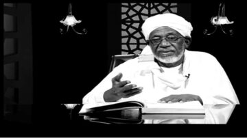 رجل مسن يرتدي عمامة بيضاء وثوب تقليدي يجلس أمام كتاب مفتوح، في خلفية داكنة مع تفاصيل إضاءة أنيقة. يتحدث بطريقة تعبيرية، مما يدل على اهتمامه بالموضوع.