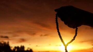 يد مرفوعة تحمل سبحة أمام غروب الشمس، مع ألوان دافئة في السماء. تعكس الصورة روحانية وتأمل.