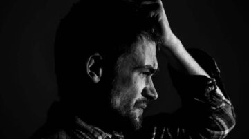 شخص يظهر في صورة بالأبيض والأسود، يعبر عن مشاعر التوتر والقلق، مع تعبير وجه حاد ويد مرفوعة على الرأس.