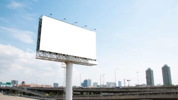 لوحة إعلانات فارغة على عمود، تقع فوق طريق سريع، مع منظر لمدينة حديثة في الخلفية وسماء زرقاء.