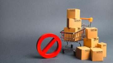 عربة تسوق صغيرة تحتوي على صناديق كرتونية، مع وجود رمز تحذيري يمنع التسوق. الخلفية رمادية، مما يبرز العناصر. الصورة تعكس فكرة عدم الشراء أو القيود التجارية.