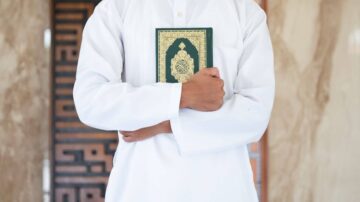 رجل يرتدي جلبابًا أبيضًا يحمل القرآن في يده، يظهر خلفه خلفية من الجدران المزخرفة.