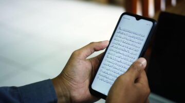 شخص يحمل هاتفًا ذكيًا مع عرض نص باللغة العربية، يبدو أنه نص ديني، في بيئة داخلية مضاءة.