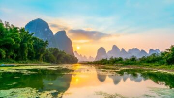 منظر طبيعي خلاب لنهر يتدفق بين جبال كارست في غوانغشي، الصين. تتلألأ أشعة الشمس عند الغروب على سطح الماء الهادئ، مع انعكاسات للجبال والأشجار المحيطة، مما يخلق مشهداً ساحراً.