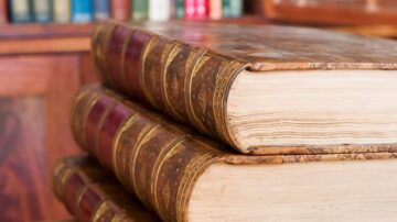 كتب قديمة ذات غلاف جلدي بني، موضوعة فوق بعضها البعض، تظهر تفاصيل حواف الصفحات. الخلفية تحتوي على مكتبة مليئة بالكتب الملونة.