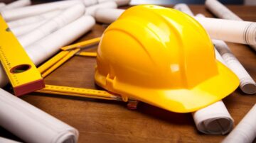 خوذة عمل صفراء على طاولة خشبية محاطة بأسطوانات خطط مع مسطرة ومسطرة زاوية.