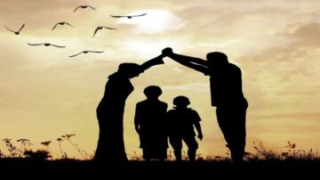 عائلة تتكون من أربعة أفراد في وضعية ظلية، حيث يقف الوالدان معًا في الخلف ويُرفع ذراعهما فوق الأطفال في مشهد يعبر عن الحماية والمحبة. السماء مليئة بالغيوم، مع ظهور طيور تحلق في الأفق.