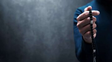 يد مرفوعة تحمل سبحة سوداء مع خلفية رمادية داكنة، تعكس روحانية وهدوء.