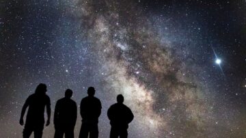 أربعة أشخاص يقفون silhouetted أمام سماء مليئة بالنجوم، مع مجرة درب التبانة تتلألأ في الخلفية. مشهد ليلي يعكس جمال الكون وسحر الفضاء.