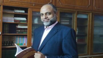 رجل يحمل كتابًا ويبتسم، يقف أمام مكتبة تحتوي على مجموعة من الكتب. يرتدي بدلة زرقاء وقميص أبيض، ويظهر خلفه رفوف خشبية مليئة بالكتب.