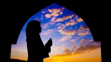 امرأة ترتدي حجابًا تصلي في ظل قوس مع غروب الشمس في الخلفية، السماء مليئة بالغيوم الملونة.