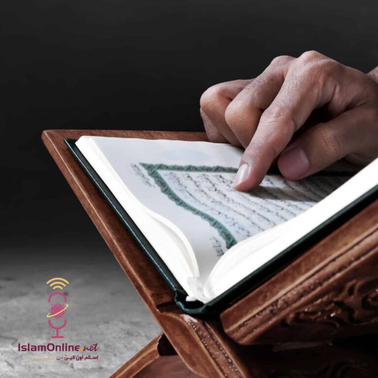 يد تظهر وهي تشير إلى صفحة من القرآن الكريم المفتوح على مصحف خشبي، مع تفاصيل دقيقة للخط العربي.