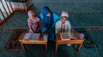 امرأة ترتدي الحجاب تجلس مع طفلين في مسجد، يدرسون القرآن. الطفلتان ترتديان أزياء تقليدية، وأمامهم مكاتب خشبية تحتوي على مصاحف وأضواء. السجاد ذو لون أخضر يضيف لمسة روحية للمشهد.